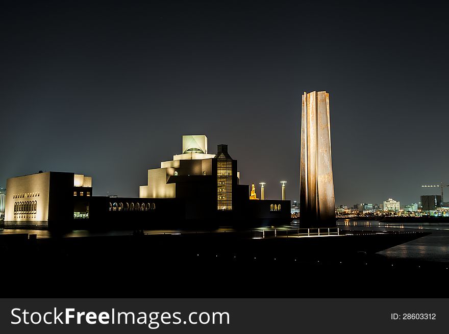 Night shot of the Doha Museum of Islamic Art, showing the strong reflections of light made by the building design and the'seven' monument on the side.