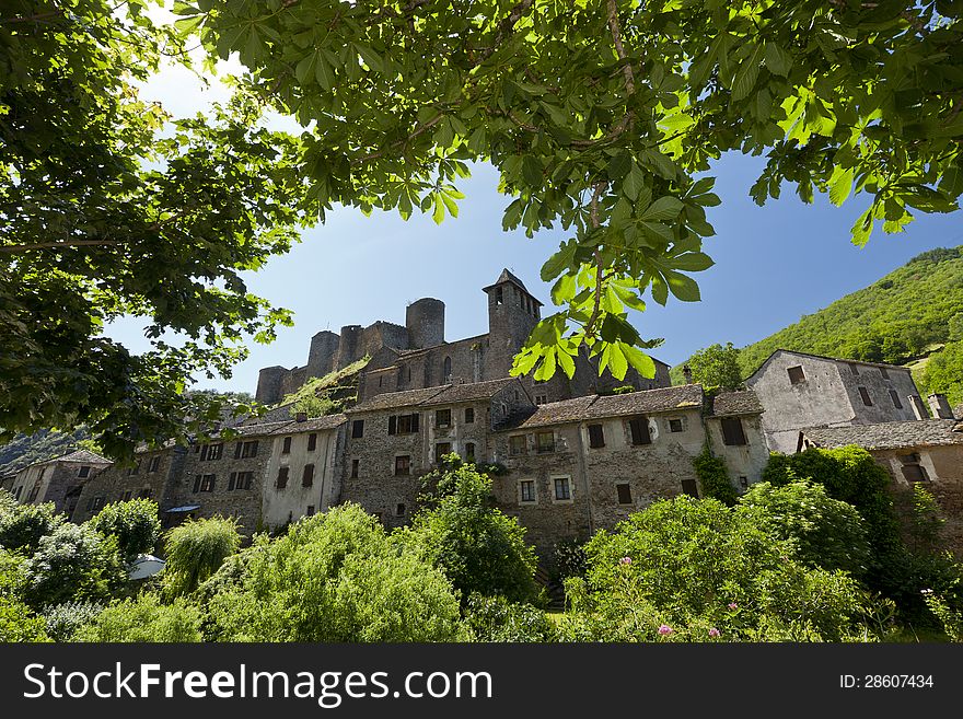 The French village of Brousse le chateau, with its castle in the background. The picture is green-framed by bushes and tree leaves.