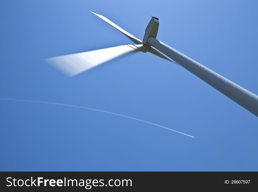 Part of a wind turbine or windmill, with in the background a curved condensation trail of an airplane.