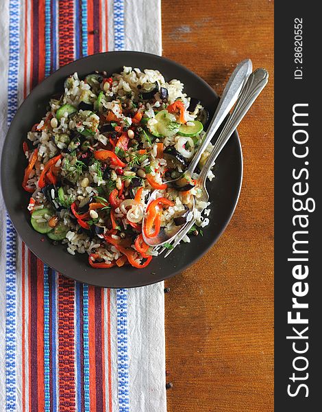 Portion of vegetarian cooked brown rice with roasted courgette, eggplant, sweet red pepper, pine nuts, peppercorns and chopped fresh dill on plate on a wooden surface