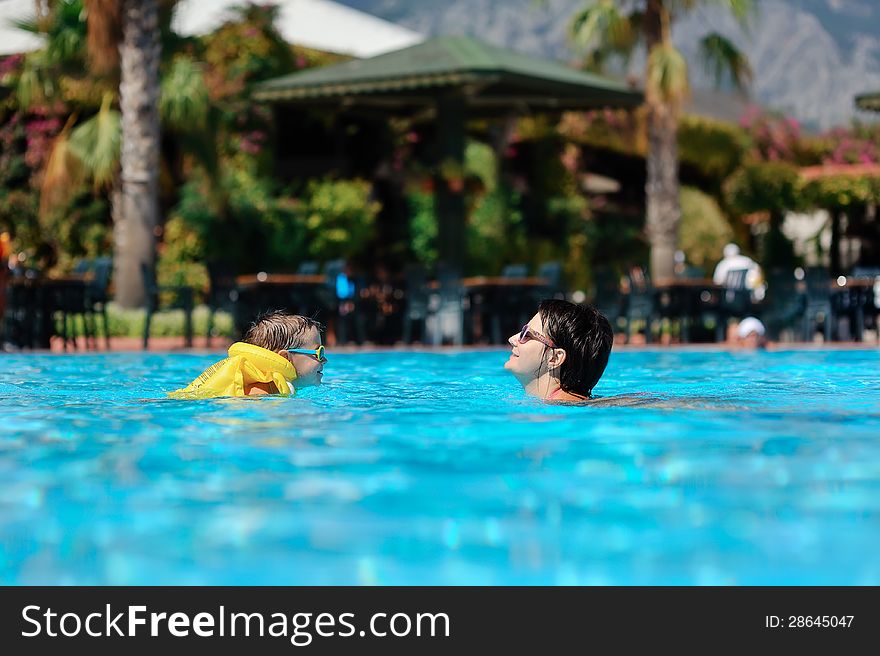 In the sunny day in the summer the boy in sun glasses and in a life jacket plays pool with mum. In the sunny day in the summer the boy in sun glasses and in a life jacket plays pool with mum