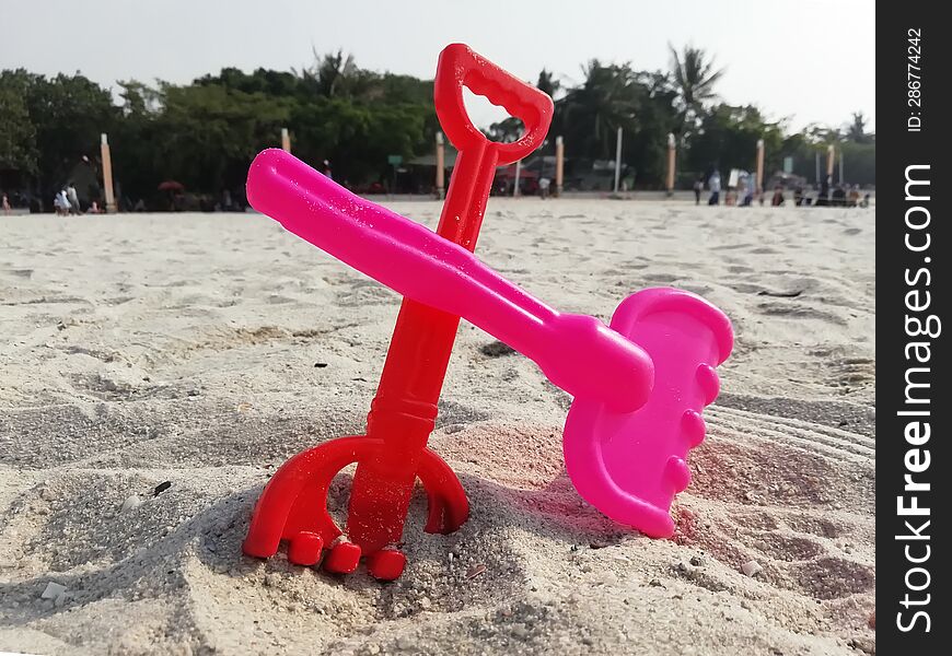 Red and pink sand rake toys in beach sand and people background taken in Jakarta Indonesia