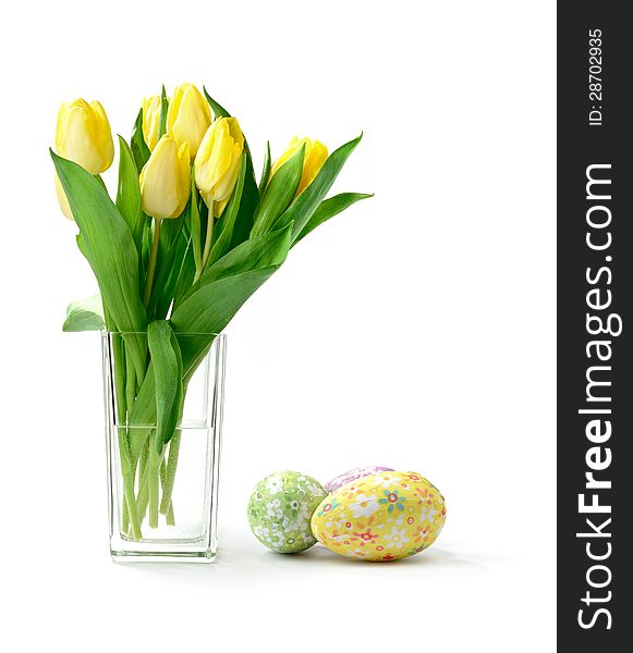 Studio macro of yellow tulips (Tulipa gesneriana) in a glass vase with decorative paper covered easter eggs against a white background. Copy space. Studio macro of yellow tulips (Tulipa gesneriana) in a glass vase with decorative paper covered easter eggs against a white background. Copy space.