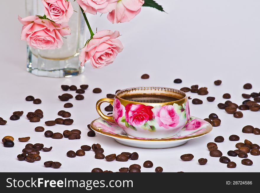 Small cup of coffee from an old service that scattered coffee beans and the background - a vase of beautiful roses. Small cup of coffee from an old service that scattered coffee beans and the background - a vase of beautiful roses