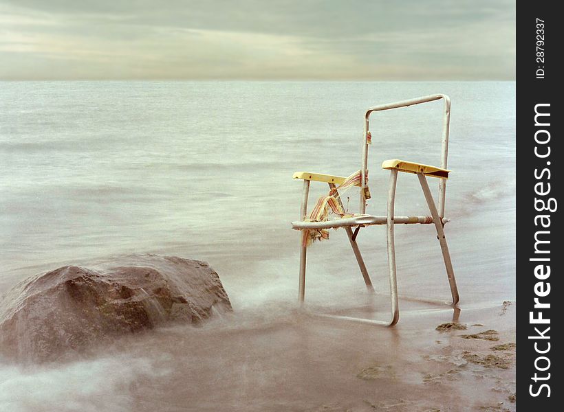 Old wrecked empty frame lawn chair sitting on the shore next to a boulder, with a cloudy sky. Old wrecked empty frame lawn chair sitting on the shore next to a boulder, with a cloudy sky.