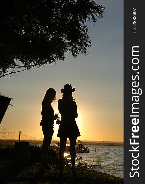 Two women having sundowners at sunset at the adge of the water. Silhouette of the women.