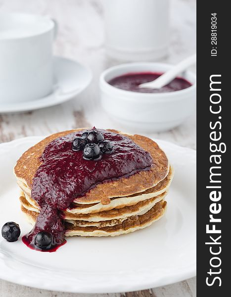 Stack of pancakes with black currant jam on a white plate, a bowl of jam in the background. Stack of pancakes with black currant jam on a white plate, a bowl of jam in the background