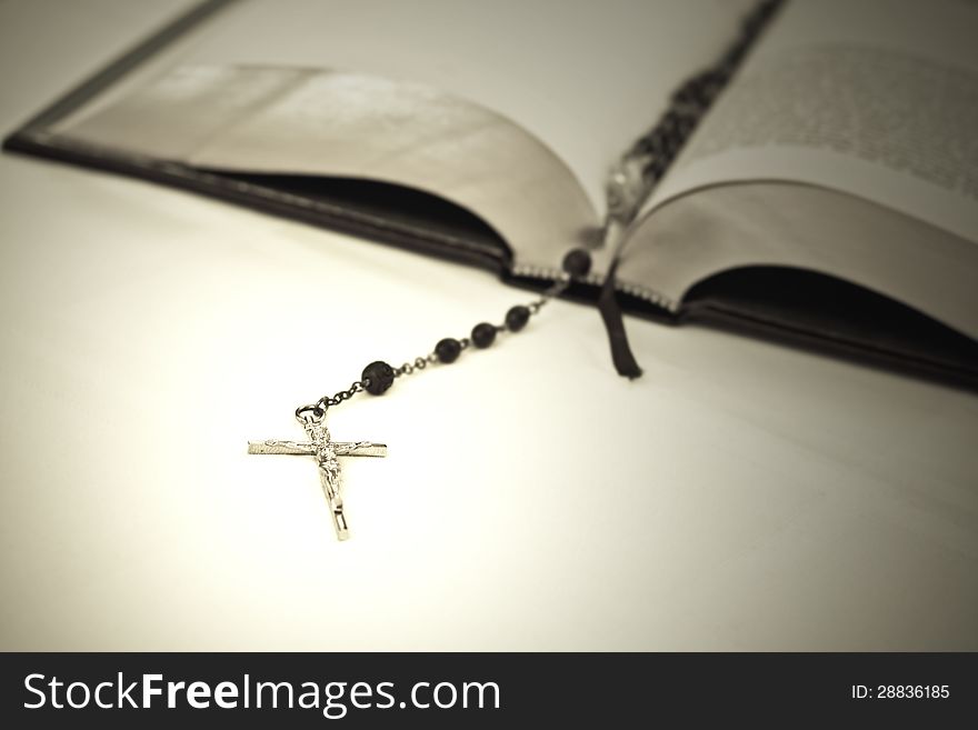 The Bible is in the background in soft focus with a rosary draped over it. The crucifix is trailing into focus in the foreground and is glowing.