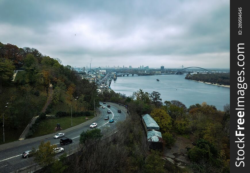 Aerial View Of Road With Passing Cars, On Hillside Against Backdrop Of River Dnieper And City Kyiv.