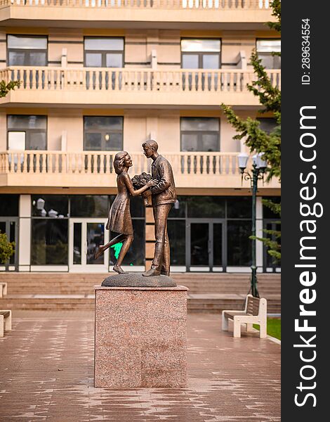 A statue of a couple of lovers, a guy gives a girl a bouquet of flowers, against the backdrop of a multi-storey building, in the a