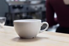 Close-up. White Low Cup On Light Wood Table Against Background Of Coffee Machine In Blur Stock Photo