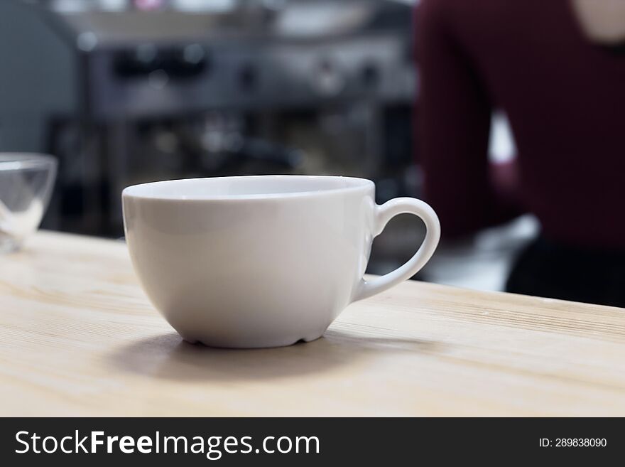 Close-up. White low cup on light wood table against background of coffee machine in blur