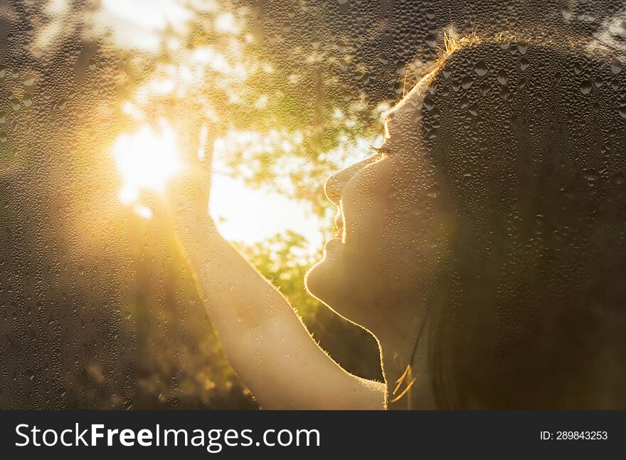 Beautiful young woman face with long dark hair on background of sunshine view through wet glass with raindrops. close-up