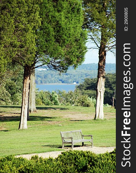 A vacant bench shownn on clear warm day in spring, summer, or early fall with trees, grass, greenry with blue water in background. A vacant bench shownn on clear warm day in spring, summer, or early fall with trees, grass, greenry with blue water in background