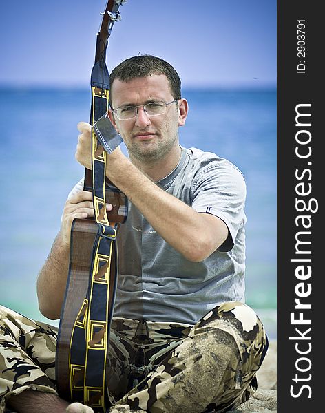 Young man wearing glasses and playing the guitar outdoors; sea and cloudy sky in the background. Young man wearing glasses and playing the guitar outdoors; sea and cloudy sky in the background.