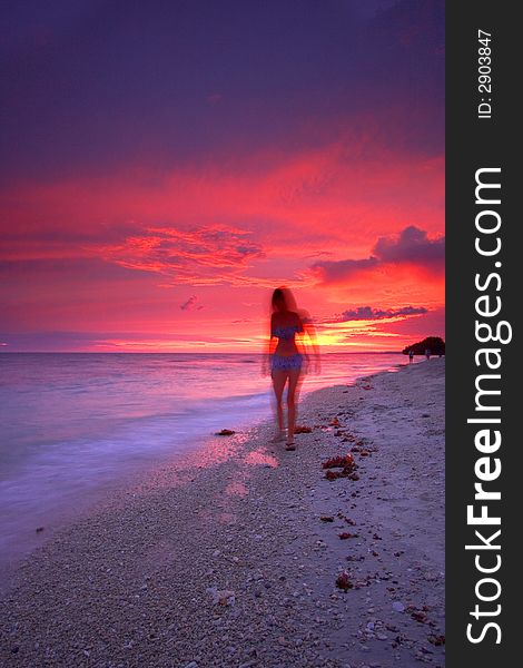An attractive single woman walking on a beautiful tropical paradise beach at sunset beside a calm sea while on a relaxing vacation. An attractive single woman walking on a beautiful tropical paradise beach at sunset beside a calm sea while on a relaxing vacation