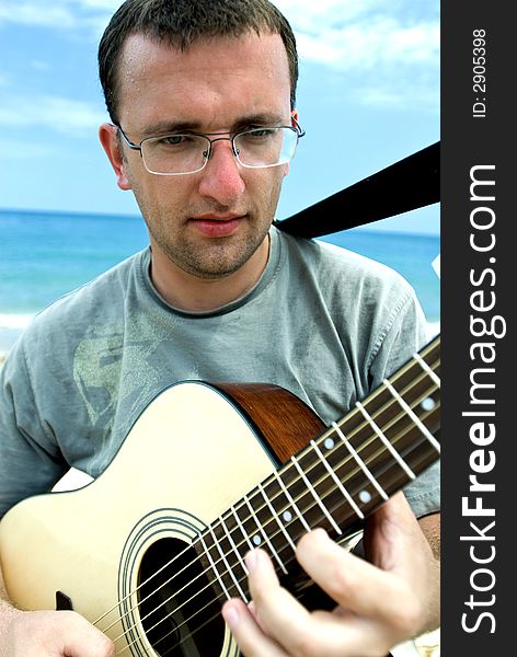 Young man wearing glasses and playing the guitar outdoors; sea and cloudy sky in the background. Young man wearing glasses and playing the guitar outdoors; sea and cloudy sky in the background.