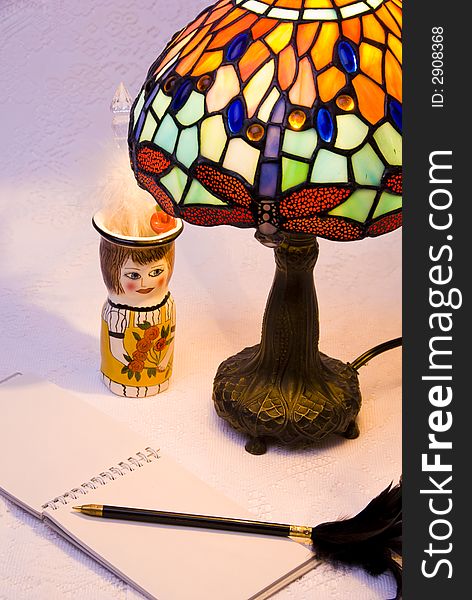 Vertical view of lit stained glass style lamp with notepad, black pen with feather and ceramic pencil holder. Vertical view of lit stained glass style lamp with notepad, black pen with feather and ceramic pencil holder