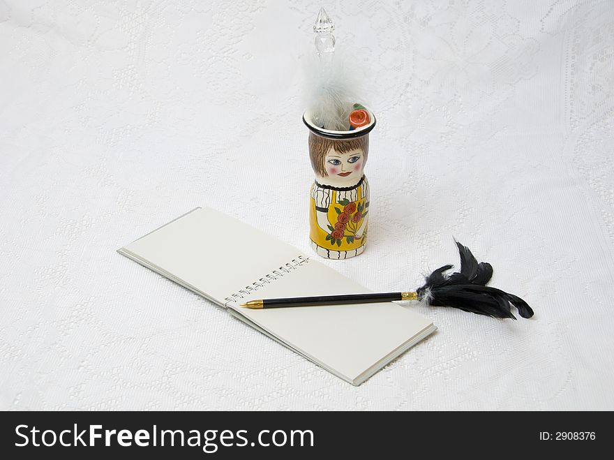 Horizontal view of ceramic pencil holder with open notebook and black pen with feathers on white lace background. Horizontal view of ceramic pencil holder with open notebook and black pen with feathers on white lace background