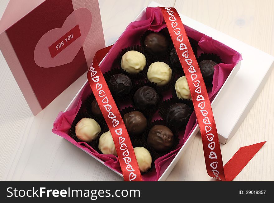 Box of chocolates with red heart ribbon and pink and red handmade gift card on white natural wood table. Box of chocolates with red heart ribbon and pink and red handmade gift card on white natural wood table.