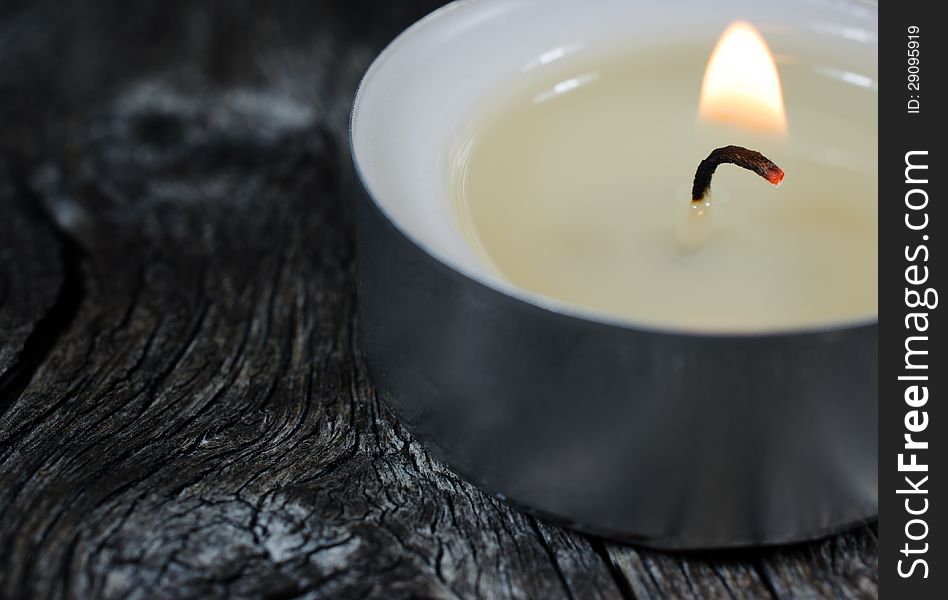 A close-up photograph of a lit tea light candle on an old wooden board with focus on the candle wick.