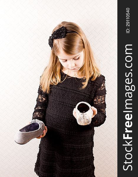 Little blonde girl standing with a different shoe in each hand making a choice of footwear to wear with her stylish black outfit isolated against a neutral background. Little blonde girl standing with a different shoe in each hand making a choice of footwear to wear with her stylish black outfit isolated against a neutral background.