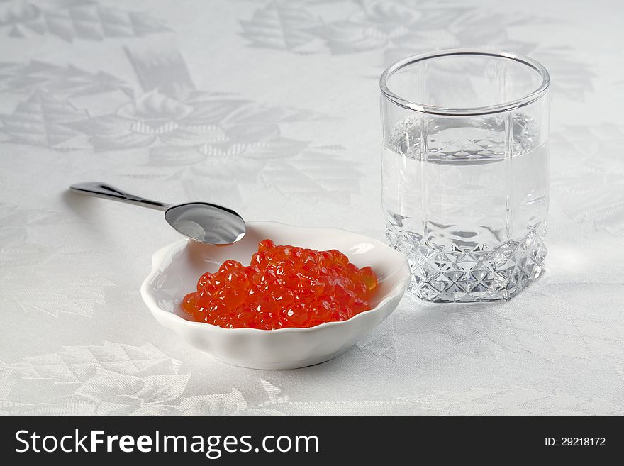 A small glass of vodka, salmon caviar in a plate for caviar and small spoon. A small glass of vodka, salmon caviar in a plate for caviar and small spoon