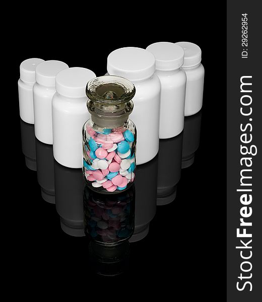 Glass bottle with multi-colored pills and row of white small bottles for pills on a black background. Glass bottle with multi-colored pills and row of white small bottles for pills on a black background.