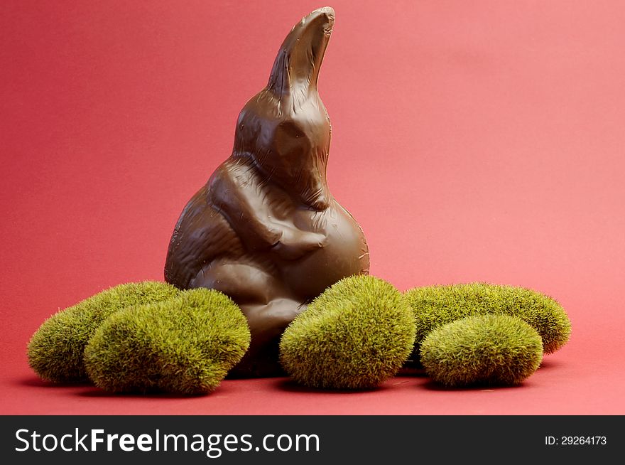 Australian alternative to the Easter bunny rabbit, a chocolate Bilby holding an egg on green moss rocks against a red background.