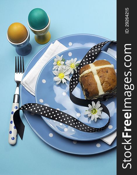 Happy Easter dinner table setting with blue polka dot plates, and decorations against a blue background. Vertical with Easter Hot Cross Bun and color boiled eggs. Happy Easter dinner table setting with blue polka dot plates, and decorations against a blue background. Vertical with Easter Hot Cross Bun and color boiled eggs.