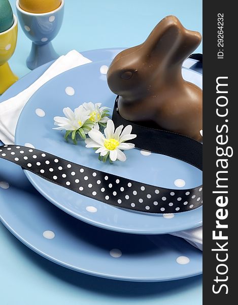 Happy Easter dinner table setting with blue polka dot plates, and decorations against a blue background. Vertical close-up with chocolate Easter bunny rabbit. Happy Easter dinner table setting with blue polka dot plates, and decorations against a blue background. Vertical close-up with chocolate Easter bunny rabbit.
