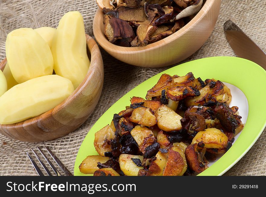 Roasted Potato with Mushrooms, Onion and Spices. Arrangement of Prepared Potato on Green Plate and Two Wood Bowls with Raw Potato and Slices of Mushrooms and Fork with Knife close up