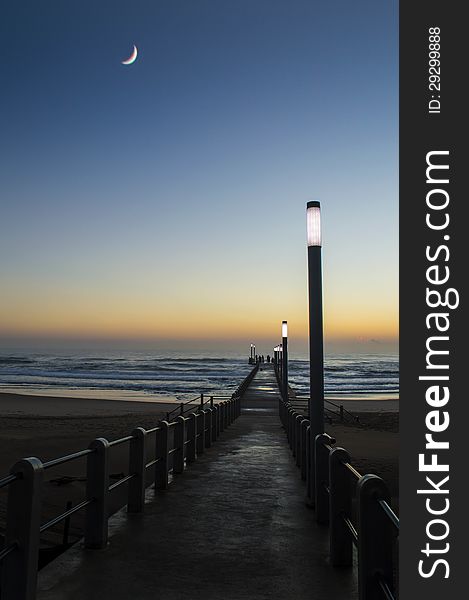 An image of a crescent moon hanging low in the sky during the early morning light over a beach with a pier still light up by light poles. An image of a crescent moon hanging low in the sky during the early morning light over a beach with a pier still light up by light poles.