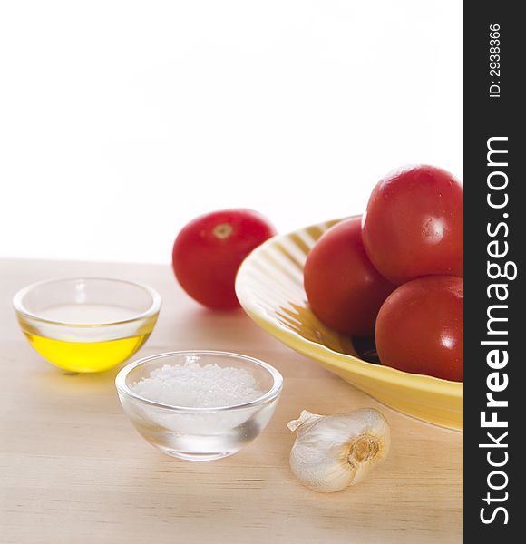 A portion of olive oil, small portion of salt, garlic clove, and a yellow plate with tomatoes on top of a wooden cutting board against a white background. A portion of olive oil, small portion of salt, garlic clove, and a yellow plate with tomatoes on top of a wooden cutting board against a white background.