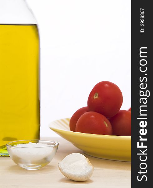 Olive oil bottle, small portion of salt, garlic clove, and a yellow plate with tomatoes on top of a wooden cutting board against a white background. Olive oil bottle, small portion of salt, garlic clove, and a yellow plate with tomatoes on top of a wooden cutting board against a white background.