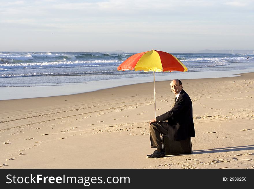 Businessman, dressed in suit and tie, relaxing under a colored umbrella watching the last rays of light go down over the beach. Businessman, dressed in suit and tie, relaxing under a colored umbrella watching the last rays of light go down over the beach