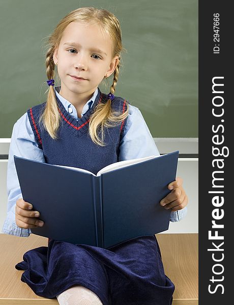 Little girl sitting on desk in front of blackboard. Holding book and smiling. Looking at camera. Front view. Little girl sitting on desk in front of blackboard. Holding book and smiling. Looking at camera. Front view