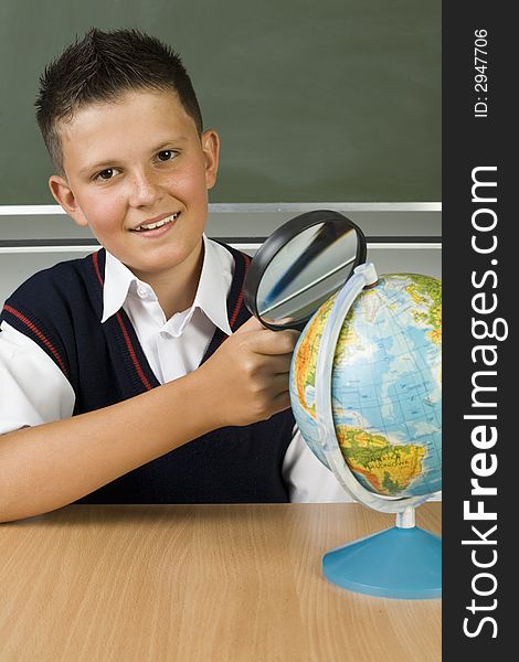 Young, smiling boy sitting at desk, nearby globe. Holding magnifying glass. Looking at camera, front view. Young, smiling boy sitting at desk, nearby globe. Holding magnifying glass. Looking at camera, front view