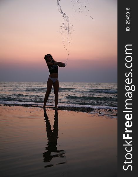 Young woman sprinkles by water on beach on sunset