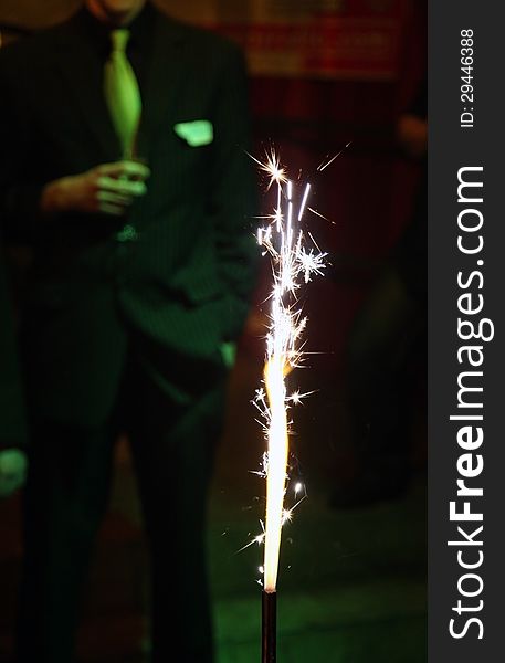 Birthday party. Focus on the sparkler in the foreground. In the background a man wear suits and hold a a glass of champagne.