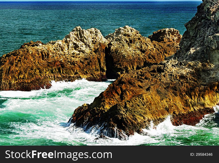 A dramatic landscape scene of a choppy sea with the waves rolling in and crashing against the huge colorful jagged rocky cliff at the point of the coast of the tropical island. Photo taken at Point Lookout, Stradbroke Island, Queensland, Australia. A dramatic landscape scene of a choppy sea with the waves rolling in and crashing against the huge colorful jagged rocky cliff at the point of the coast of the tropical island. Photo taken at Point Lookout, Stradbroke Island, Queensland, Australia.