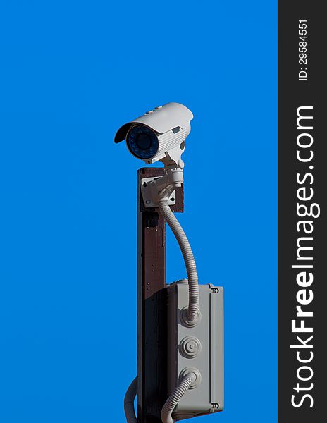 A surveillance camera installed on a pole, watching everything that moves within its covering radius. Shot against the blue sky as background. A surveillance camera installed on a pole, watching everything that moves within its covering radius. Shot against the blue sky as background