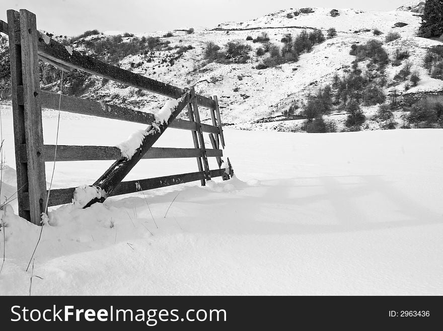 A monochrome image of a snowy gate opeing onto a snow covered field