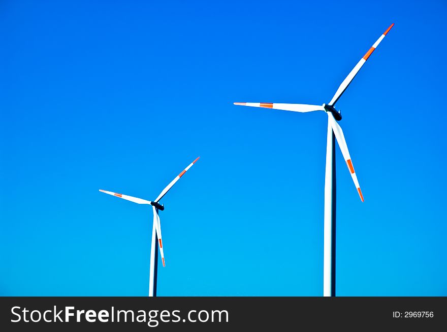 Two electricity generating windmills against a blue sky. Two electricity generating windmills against a blue sky.