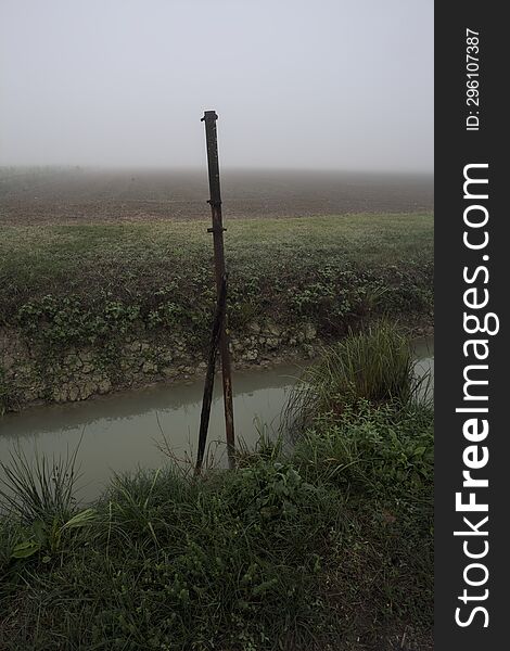 Pole by the edge of a ditch with water on a foggy day in the italian countryside