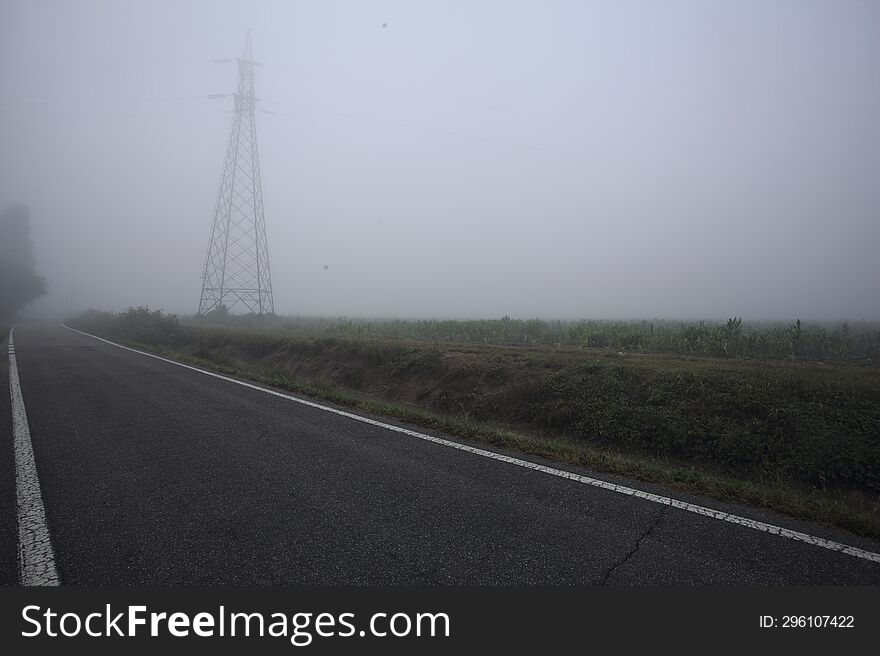 Road bordered by trenches with a pylon in a field by its edge on a foggy day in the italian countryside