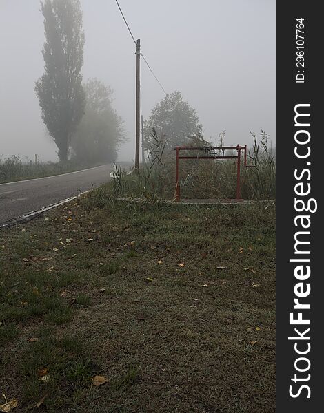 Narrow Road Bordered By A Few Trees And Trenches With Weirs On A Foggy Day In The Italian Countryside