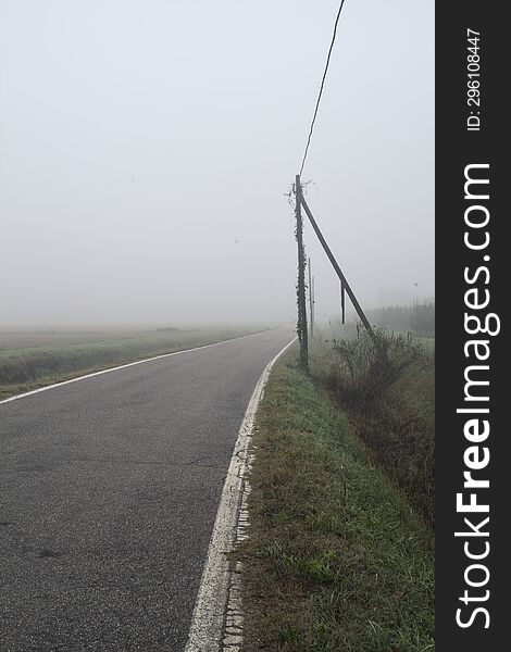 Road bordered by a trench and a power line next to fields on a foggy day in the italian countryside