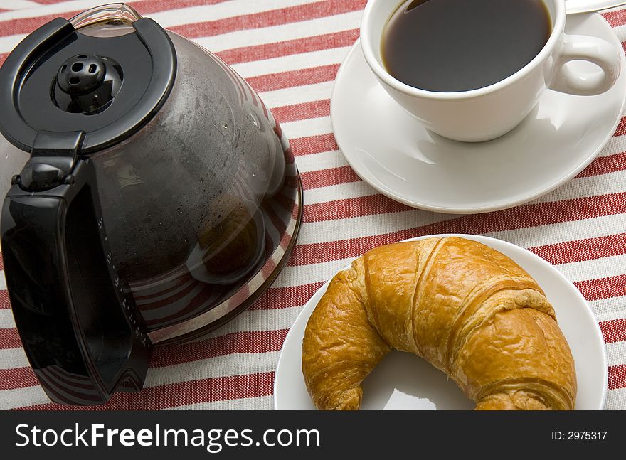 A pot of freshly brewed coffee, a cup of black coffee and a croissant on a plate on a breakfast table with red and white pattern fabric table cover. A pot of freshly brewed coffee, a cup of black coffee and a croissant on a plate on a breakfast table with red and white pattern fabric table cover.