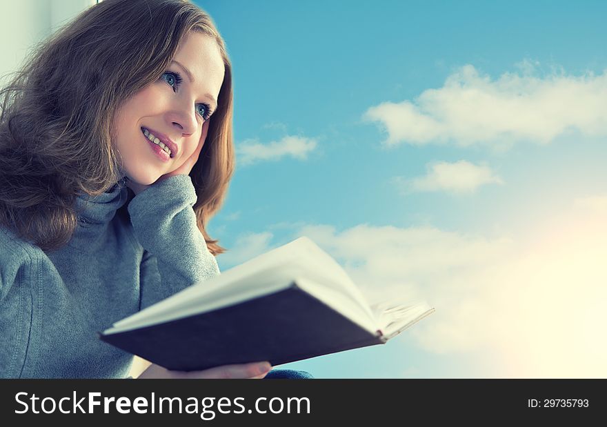 Beautiful young woman reading a book while sitting at a window in the sky and sunset